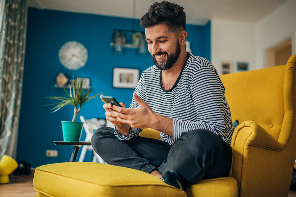 Man sits on a yellow chair in a modern and room smiling qt his phone in his hand
